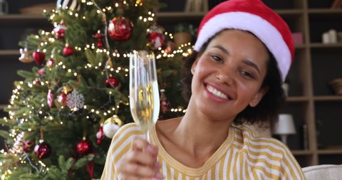 Happy New Year everyone. Smiling teen Black female in Xmas hat wave hand to family from screen say best wishes toast drink champagne. Excited young woman greet friends with Christmas remotely