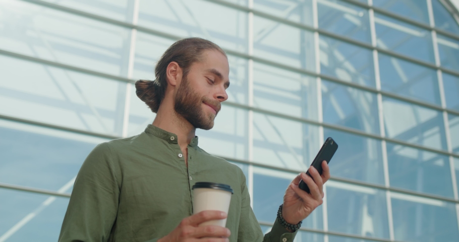 Happy Businessman standing near Office Building Messaging using Smartphone. Holding a cup of coffee looking at Phone Screen while typing Message. Smiling Young Caucasian Man. Device User. Apps. Royalty-Free Stock Footage #1081629206