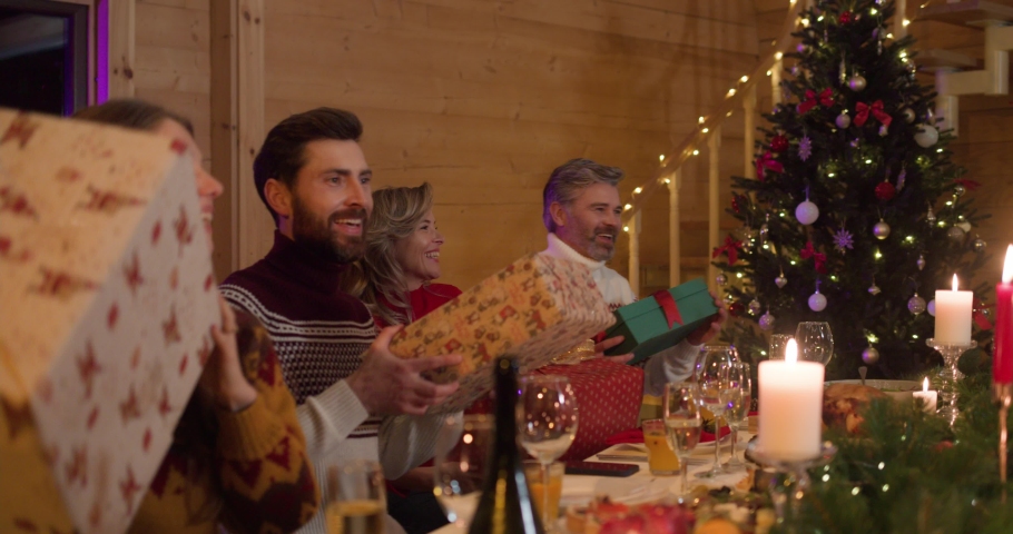 Happy Family Exchanging Presents during Christmas Dinner at Home. Big Family enjoying Winter Holidays and Gifts Together standing near Christmas table. Happy Relatives Gathering. Greeting. Hugging. | Shutterstock HD Video #1081629326