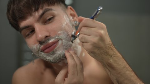 Close-up front view man shaving cheek smiling in slow motion. Portrait of confident Caucasian guy shaving off stubble in bathroom at home indoors. Routine and hygiene concept