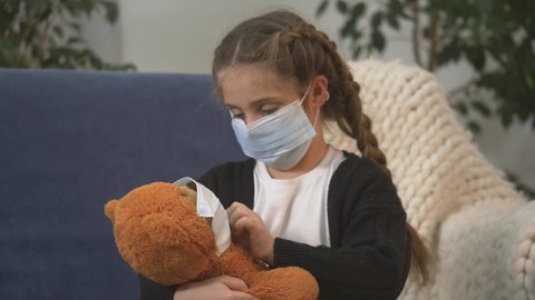 little girl medical mask sits at home in quarantined living room isolation. girl hugs teddy bear, plays with her favorite toy. child hugs bear showing love,care in family. child in medical mask.