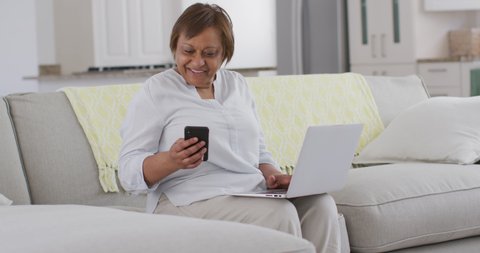 Happy african american senior woman sitting on couch using smartphone and laptop, smiling. retirement lifestyle, leisure time alone at home with technology.