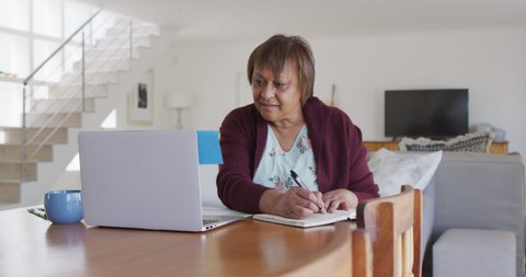 Smiling african american senior woman sitting at dining table, using laptop and writing in notebook. retirement lifestyle, at home with technology.