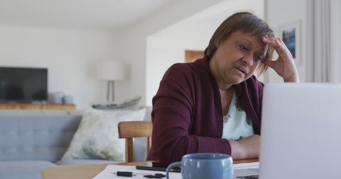 Worried african american senior woman at dining table, using laptop and holding head. retirement lifestyle, at home with technology.