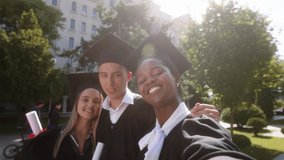 Group of multiracial students graduates two ladies and one guy taking selfie video with the camera smiling large after the graduation