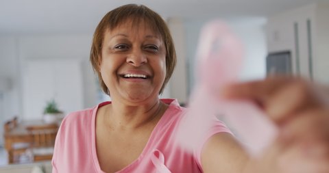 Smiling african american senior woman wearing pink t shirt holding pink breast cancer ribbon. senior female lifestyle and breast cancer positive awareness campaign.