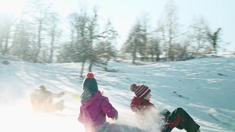 Tracking shot of a smiling Caucasian children sledding delighted with plastic shovel sleds and enjoying the pleasure of sledding and snow
