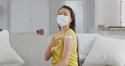 Happy asian woman in face mask sitting on sofa showing arm with plaster after vaccination. health and lifestyle during covid 19 pandemic.