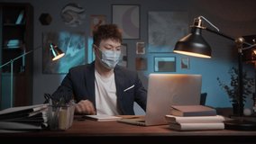 Young Asian man wearing medical mask finishing working remotely at home during coronavirus lockdown. Korean student, Chinese guy using video call lecture for remote learning.