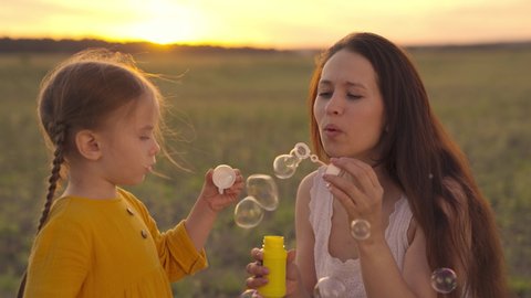 Happy Mother and her little Daughter playing together outdoor, blowing soap bubbles, having fun on backyard. Nature. Beauty Mum and her Child in Park together. Family, Mother's Day Joy. Mom and Baby