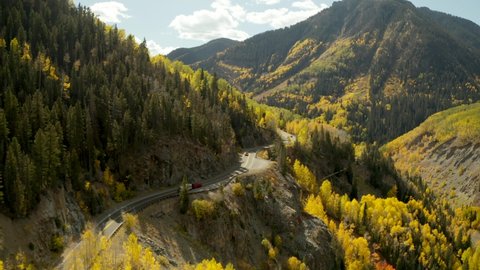 An aerial drone flight following a truck with RV trailer driving up a winding road through the Rocky Mountains of Colorado during the colorful autumn season.