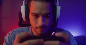 Happy Asian man gamer wear headphone competition play video game online with smartphone colorful neon light in living room at night modern house. Esport streaming game online, Home quarantine activity.