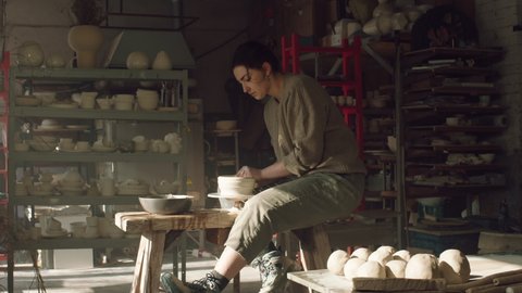 Woman potter is sitting on potter's wheel and making a bowl of wet clay, working with inspiration in beautiful workshop, enjoying pottery craft, Slow motion.