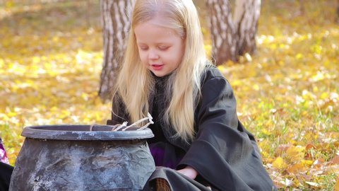 A little girl in a black mantle takes out of the cauldron toy skeletons of the gallows sitting against the background of an autumn park. Children's Halloween.