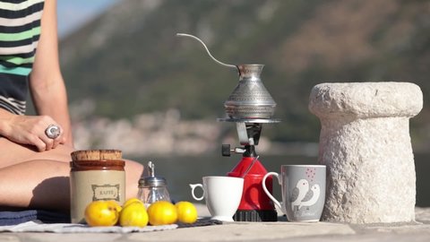 Unrecognizable woman sits on ground at picnic in Montenegro and make coffee in cezve on camp gas burner at pier in bay of Donji Stoliv. Woman make coffee and then go away