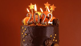 Happy New Year's Eve 2022 chocolate cake decorated with gold burning candles on against dark wood table setting background. Close up cinemagraph loop static.