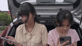 Two Asian adorable teen girls enjoy watching social video online on mobile smartphone with earphones with sitting together in rear side of hatchback car. Young lifestyle. Happy Thai sibling.