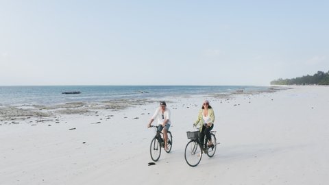 4K real-time drone footage couple of tourists riding bicycles on sandy beach on clear warm morning in Zanzibar, Tanzania. Travel concept, free time, tourist resort concept.