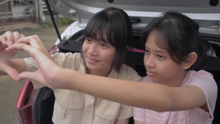 Two Asian adorable female teenagers enjoy making hand gesture heart shape together while sitting on rear side of hatchback car. Young lifestyle. Bonding relationship in sibling family.