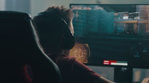 Teen gamer in headset shooting enemies during cybersport match while sitting at table and playing videogame in weekend at home