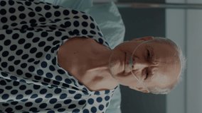 Vertical video: Portrait of elder patient with nasal oxygen tube standing on bed in hospital ward at medical clinic. Sick old man with oximeter connected at heart rate monitor to check progress