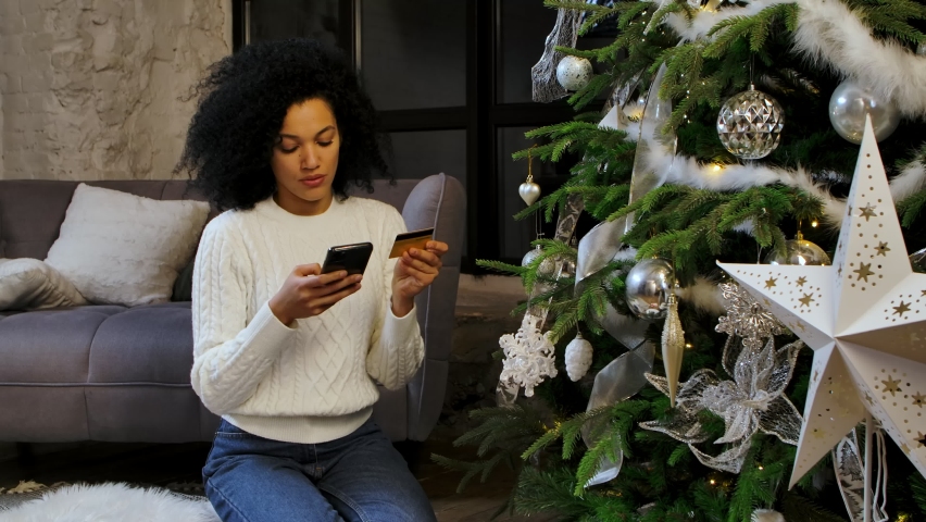 Portrait of a happy African American woman making an online purchase using her smartphone and credit card. Young woman sits near a festive Christmas tree in a bright loft style room. Slow motion.