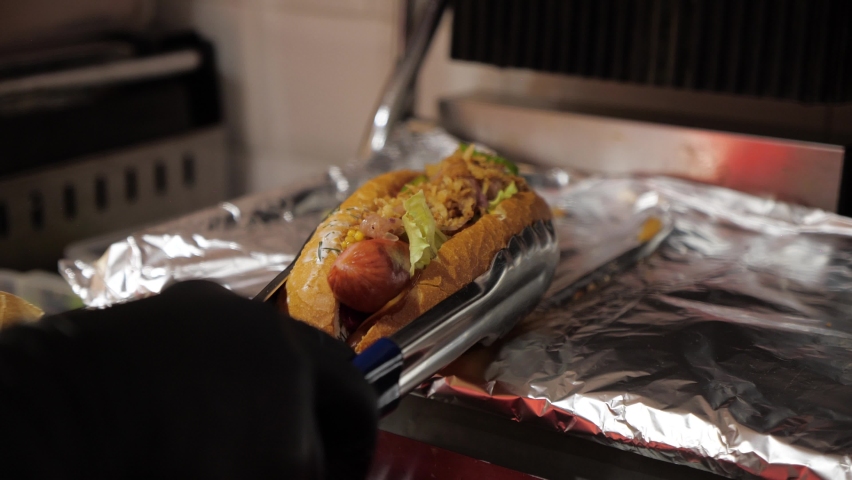 Street food in the big city. the chef removes a large, juicy hot dog from the grill and places it on a plate to serve to the guest | Shutterstock HD Video #1081669994