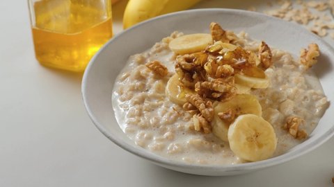 Pouring honey in oatmel porridge with banana, nuts in a plate. Healthy breakfast. Diet food. 4k video