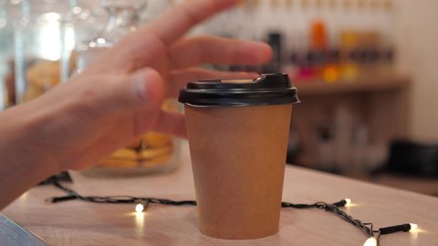 close-up in a coffee shop a man's hand picks up a cup of morning coffee to go  from the bar. yellow lights garlands and cookies in glass jars on the background. disposable cup with plastic lid