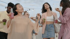 Attractive asian brunette in stylish outfit dancing actively with her diverse friends on roof top. Group of young men and women enjoying summer party with drinks and food. - Powered by Shutterstock - Get 15% off with code: PIKWIZARD15