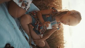 A cute preschool girl sits cross-legged and drinking milk through a straw at a family picnic in a wheat field. Vertical video. Slow motion