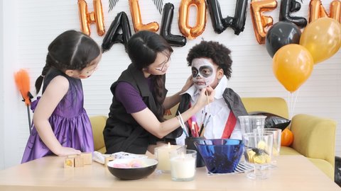 Young woman make-up artist painting vampire face  black skined child for halloween party with little girl in witch dressed.