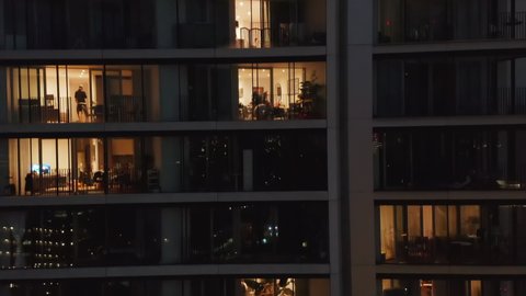 Crane up footage of high rise residential building at night. Close up view of wall with windows. People moving inside flats. London, UK