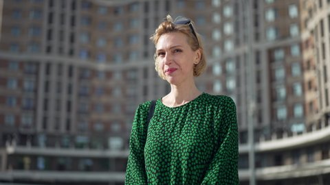 Portrait of shy Caucasian adult woman in green dress standing outdoors in sunlight smiling looking at camera gesturing. Happy smiling lady posing in urban city in slow motion. Live camera zoom in
