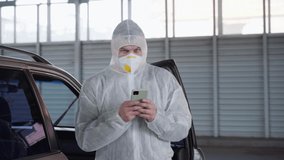 Man medical worker doctor dressed white protective overalls standing car on parking resting between work using smartphone during covid pandemic. Candid shoot male person protective suit take a break 