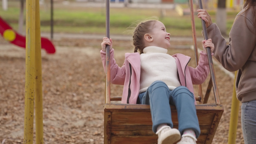 little kid swinging on a swing and laughing while flying up, an autumn playground, a happy family, a mother rolls child in city park, childhood dream to fly, smile at mom on a walk, play with a baby Royalty-Free Stock Footage #1081701140