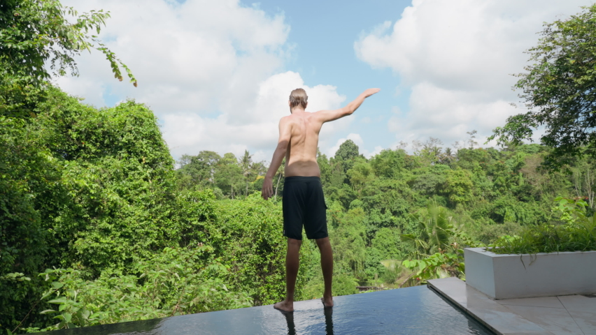 A tall athletic young man in black shorts stands on the edge of the pool and does a morning workout. A man does exercises waving his arms on the edge of the pool with a view of jungle. Slow motion.