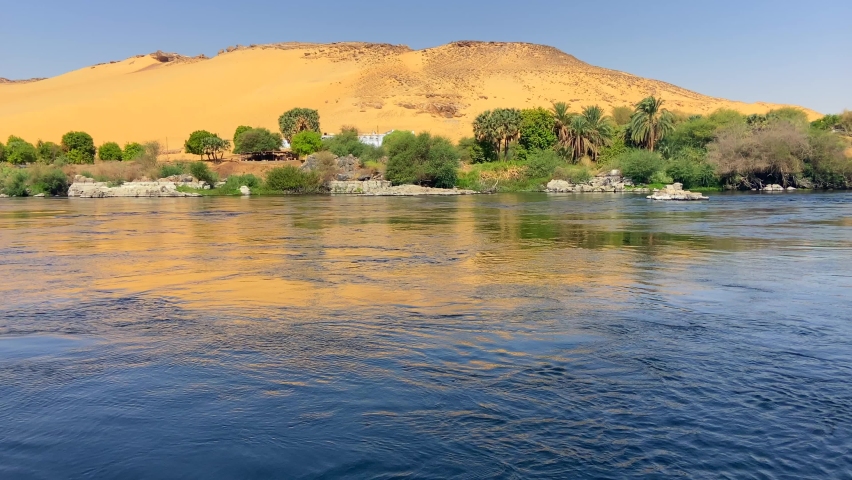Little oasis landscape on the banks of the Nile with a huge dune with many green vegetation at its base. 4K footage of a cruise sailing through Nile river. Concepts of Egyptian tourism and travel. Royalty-Free Stock Footage #1081704089