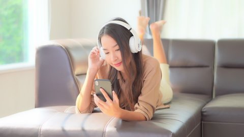 A young woman playfully kicks her feet while lying on the couch as she listens to music through her smartphone and headphones.
