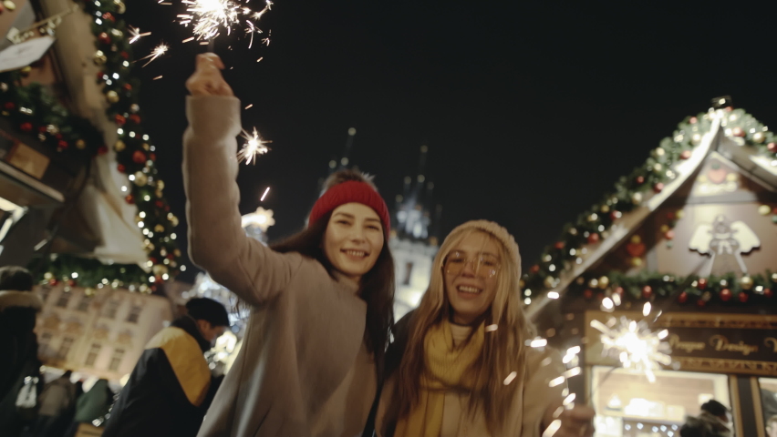 Smiling women walking a festive fair on Christmas Eve in New York. Pretty young girlfriends laughing and dancing with sparklers in street. Being happy together and making dreams come true in Christmas