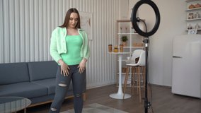 Young pretty girl dancing at camera using smartphone on tripod and ring lamp at home, makes trendy content on a mobile app to share on social media.