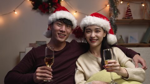 love romance holiday celebration concept. engaged couple with red santa hat holding and drinking wine glasses sitting on sofa during video call online. joy female and male toasting with white wine