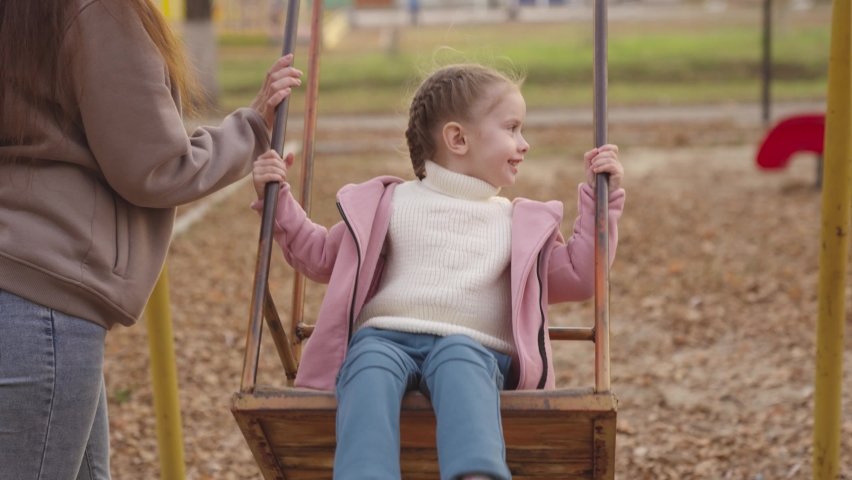 mother shakes little child on playground swing, cheerful kid flies up and down, baby laughs and smiles while playing, mother and daughter on walk in city park, happy family, childhood dream of flying Royalty-Free Stock Footage #1081723328