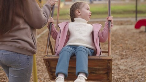 mother shakes little child on playground swing, cheerful kid flies up and down, baby laughs and smiles while playing, mother and daughter on walk in city park, happy family, childhood dream of flying