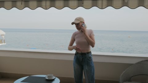 A woman in a baseball cap, nervously talking on the phone on the balcony of a hotel room overlooking the sea.