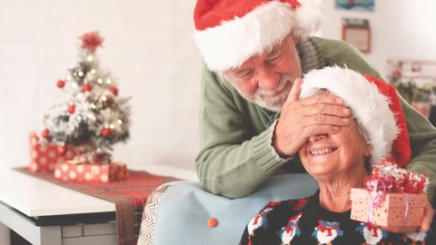 Senior couple in warm clothing and santa hat, man covering eyes of old wife with a surprise gift for her. Loving old romantic heterosexual couple celebrating christmas festival together
