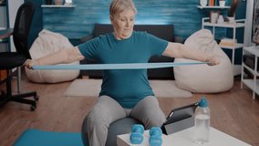 Old woman watching online workout program on tablet, sitting on toning ball. Retired person pulling resistance band to stretch arms muscles while following training lesson on video.