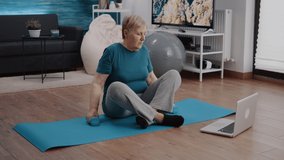 Aged person lifting weights with dumbbells and watching video of training lesson with coach. Elder woman doing physicial exercise to train arm muscles while following online workout program
