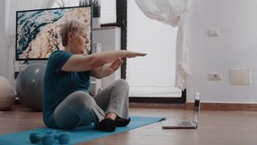 Senior woman following training video program on laptop to do workout and physical activity. Retired person watching online lesson with trainer to exercise fitness and gymnastics on mat.
