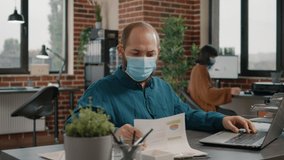 Business man doing paperwork and looking at rate charts while working with laptop. Employee wearing face mask and analyzing data analysis on files for project planning and strategy. - Powered by Shutterstock - Get 15% off with code: PIKWIZARD15